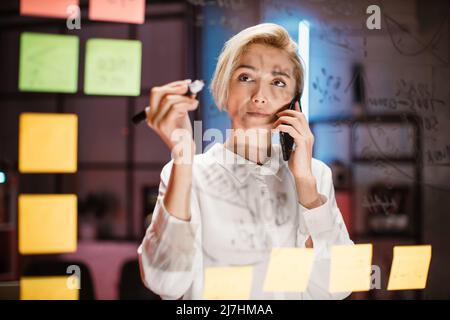 Strategy, business and scrum kanban concept. Confident experienced Caucasian female manager or ceo holding marker and writing on glass board in office room, while talking with colleague on phone Stock Photo