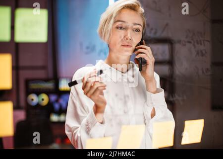 Business woman writing on transparent glass wall with sticky notes in office. Confident focused blond lady, discussing business ideas and plans talking on the phone with colleague Stock Photo