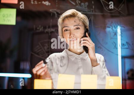 Happy smiling blond woman working in office, brainstorming near the glass board with sticky notes, writing new ideas and talking with colleague using smartphone Stock Photo