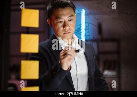 Skilled professional scrum master managing project tasks in modern evening office. Close up of concentrated asian businessman in formal wear writing notes, charts and working tasks on glass wall. Stock Photo