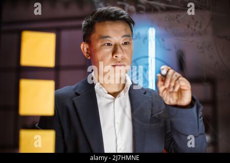 Skilled professional scrum master managing project tasks in modern evening office. Close up of concentrated asian businessman in formal wear writing notes, charts and working tasks on glass wall. Stock Photo