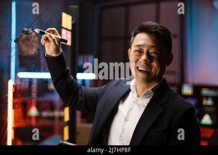 Waist-up portrait of smiling handsome Asian businessman ceo manager in formal wear, standing at glass board and writing project tasks. Night office blurred background Stock Photo
