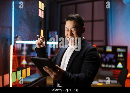 Overjoyed mature Asian businessman, standing near glass board with sticky notes, looking at the tablet pc screen and celebrating win victory of finished project. Good work results concept Stock Photo