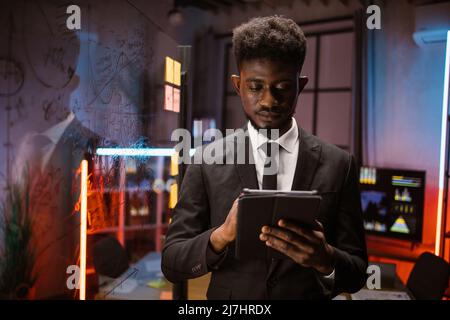 Serious african american businessman managing project on digital tablet pc, working in night office with scrum glass board. Focused male professional planning corporate strategy using computer Stock Photo