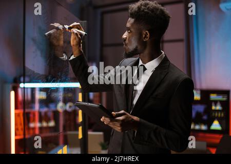Focused male african american ceo entrepreneur finance expert, using tablet while checking work plan, looking at scrum glass board with post it sticky notes in office, thinking on project management Stock Photo