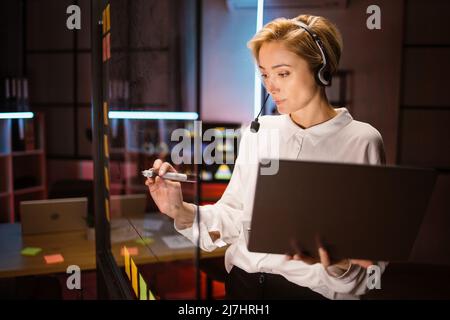 Side view of focused female worker, wearing headset and using laptop computer, writing ideas and project tasks on glass wall at dark office. Overworking process at office concept Stock Photo