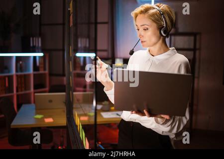 Side view of focused female worker, wearing headset and using laptop computer, writing ideas and project tasks on glass wall at dark office. Overworking process at office concept Stock Photo