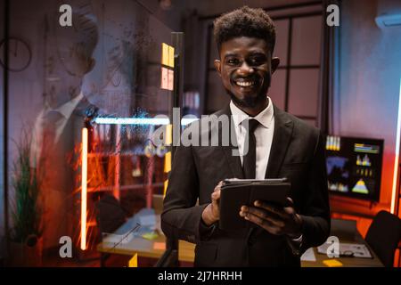 Serious african american businessman managing project on digital tablet pc, working in night office with scrum glass board. Focused male professional planning corporate strategy using computer Stock Photo