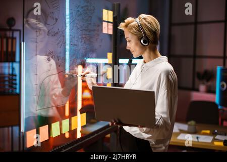 Female financial expert, ceo manager working near scrum kanban glass wall and writing ideas. Blond woman in headset, using laptop and consulting with colleague on video call Stock Photo