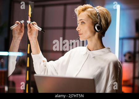 Close up side view of confident blond young businesswoman wearing white shirt and headset, writing down plans and ideas on a glass office panel while holding in her hands laptop Stock Photo