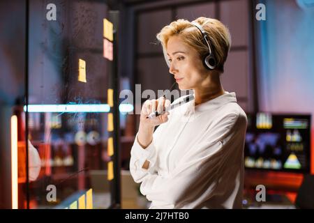 Pensive smart blond businesslady in headset, holding marker in hands and looking at the transparent glass board, while working late at dark office. Business, brainstorming, planning concept Stock Photo