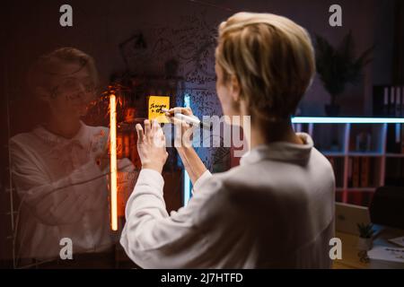 Close up rear view of business woman in white shirt, standing in front of glass kanban board, and writing ideas and tasks on yellow sticky note, developing strategy and analizing project progress. Stock Photo