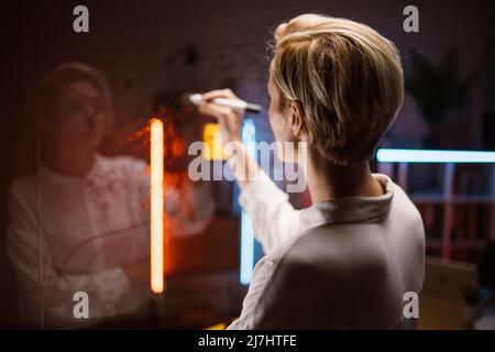 Close up rear view of business woman in white shirt, standing in front of glass kanban board, and writing ideas and tasks, developing strategy and analizing project progress. Stock Photo