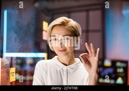 Close up head shot of pretty smiling blond Caucasian business woman in white shirt, posing on camera, while standing in modern dark office. Blurred office interior background, glass board Stock Photo
