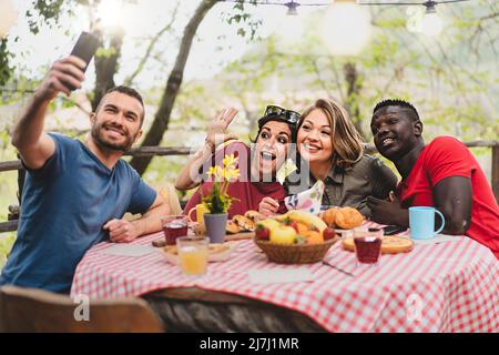 Multiracial friends relaxing taking selfie at picnic table - Young people having fun together celebrating, eating brunch outdoors at the countryside t Stock Photo
