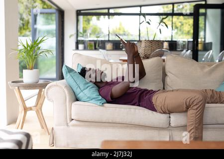 African american young man surfing net over digital tablet while relaxing on sofa in living room Stock Photo