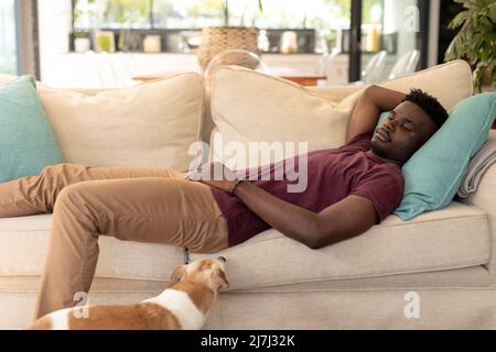 Dog looking at thoughtful serious african american young man relaxing on sofa in living room at home Stock Photo