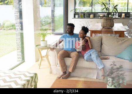 Smiling african american young couple looking through window while relaxing on sofa in living room Stock Photo