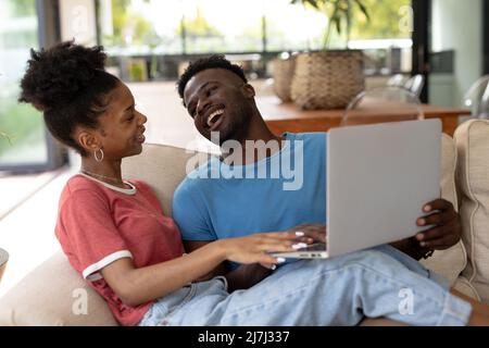Happy african american young man laughing while using laptop with girlfriend on sofa in living room Stock Photo