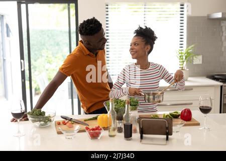 Smiling african american young man looking at girlfriend preparing food in kitchen at home Stock Photo