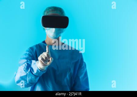 Unrecognizable person dressed in a blue medical uniform using a virtual reality headset pointing with her finger at a camera. Studio shot on blue background, focus on the foreground. High quality photo Stock Photo