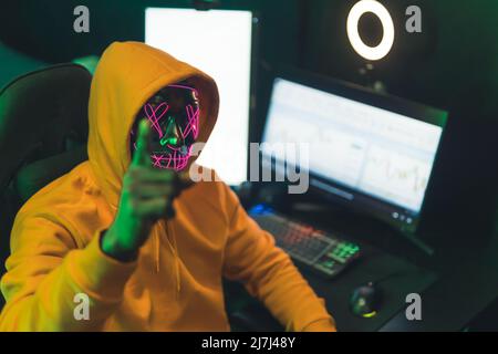 man with a scary neon mask sitting in front of a computer hacking concept medium closeup. High quality photo Stock Photo