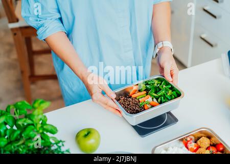 Healthy diet plan for weight loss, daily ready meal menu. Close up Woman weighing lunch box cooked in advance,ready to eat on kitchen scale. Balanced Stock Photo