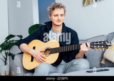 Man taking online guitar lessons via a mobile application on his phone at home sitting on the gray couch, sofa in the living room. He watching acousti Stock Photo