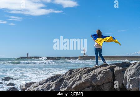Refugee woman holding national flag of Ukraine stands by the ocean. Stock Photo