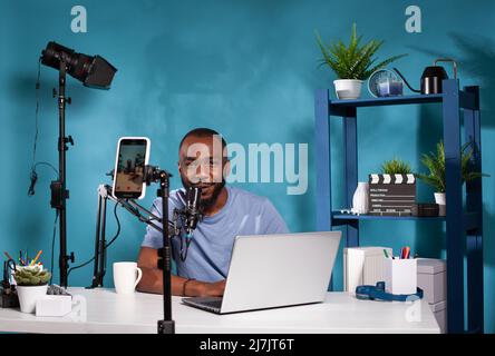 Smiling influencer interacting with audience in front of recording smartphone during online live show using laptop. Content creator talking with fanbase looking at live video podcast setup. Stock Photo