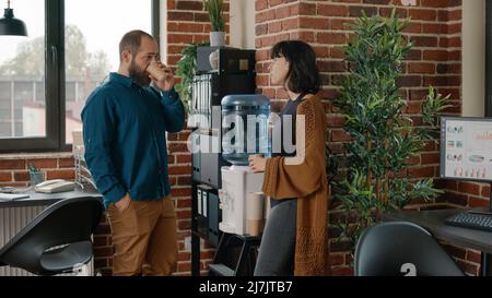 Colleagues on break having discussion and using water cooler in business office. Man and woman talking while drinking cup of water as refreshment, using liquid dispenser for hydration Stock Photo