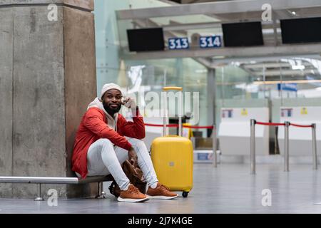 Joyful African American man sitting in airport terminal with yellow suitcase near check-in counters. Stock Photo