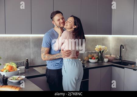 Cheerful just-married young Caucasian couple in hugs having fun and laughing in kitchen. Stock Photo