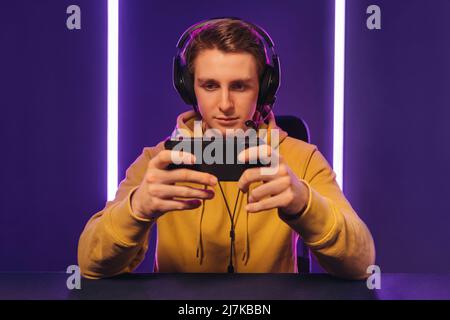 Portrait of young handsome cyber sportsman wearing headset playing online video game on his smartphone, looking at cellphone screen, focused on game, streaming. Playing mobile games, cyber sport Stock Photo