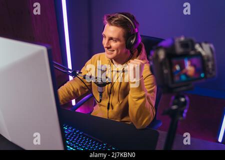 Happy guy pro gamer has won the battle, celebrating his victory, recording game stream on camera. Cyber sportsman blogger influencer shooting video while playing computer games in PC at home at night Stock Photo