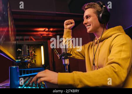 Excited young man pro gamer in headphones making winner hand gesture while playing video game on computer at home. Caucasian cyber sportsman streaming while playing in online shooter game, he just won Stock Photo