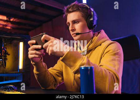 Portrait of young guy pro gamer wearing headset streaming while playing mobile video game on his smartphone in neon coloured room at home. Focused streamer participates in online e-sport tournament Stock Photo