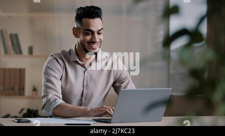 Smiling happy arab man worker businessman finished task computer work relax sit at workplace desk put hands behind head feel satisfied with work well Stock Photo