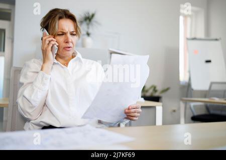 Middle aged business woman with a serious face talking on a smartphone while looking at documents Stock Photo