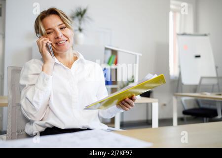 Positive female office worker with clipboard and documents in her hand talking on the smartphone Stock Photo