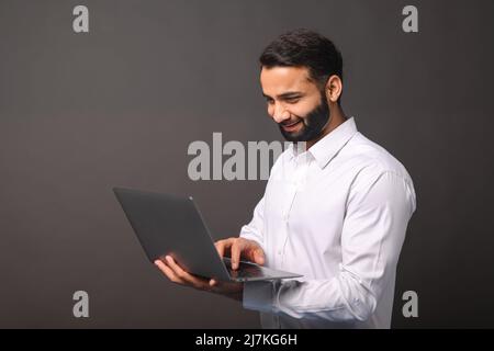 Portrait of happy young bearded Indian man in formal white shirt holding and using laptop computer isolated on black background. Male office employee typing, answering email, copy space Stock Photo