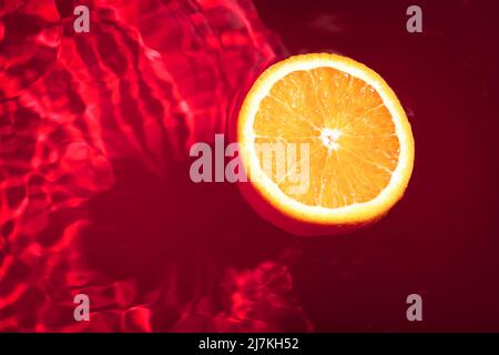 Piece of fresh orange on water surface with waves and reflections. Swimming piece of orange on red abstract background. Stock Photo