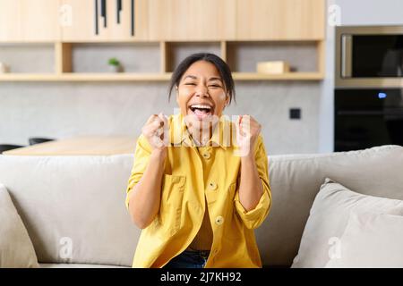 Amazed African-American lady raising hands up yelling yes loud, excited surprised dark skin lady showing win gesture sitting on the sofa at home, looking at the camera with eyes wide opened Stock Photo