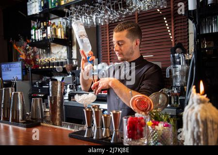 Positive young male bartender with trendy hairstyle in uniform pouring vodka into jigger while standing at counter and preparing alcohol cocktail with Stock Photo