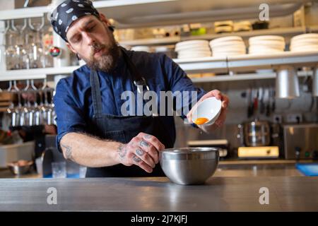 Focused bearded male chef in uniform adding raw egg into bowl while working at table in light kitchen of restaurant Stock Photo