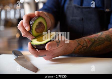 Cropped unrecognizable focused male chef in apron cutting ripe green avocado at table with cutting board while working in kitchen Stock Photo