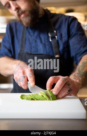 Cropped unrecognizable focused male chef in apron cutting ripe green avocado with knife at table with cutting board while working in kitchen Stock Photo