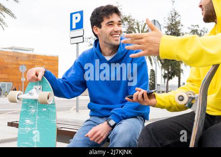Two friends skaters with colorful outfits talking to each other while sitting on a bench in street Stock Photo