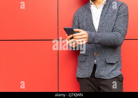 Cropped unrecognizable male entrepreneur in formal wear texting message on cellphone while standing on red background in city Stock Photo
