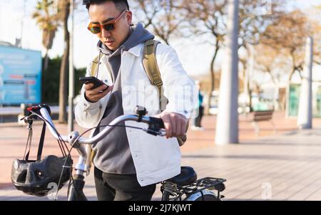 Focused male in sunglasses with bicycle texting message on cellphone while standing on paved walkway in city with tall trees Stock Photo